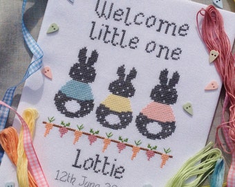 Può includere: Ricamo a punto croce con tre conigli grigi che indossano abiti di diversi colori. I conigli sono circondati da una ghirlanda di foglie verdi e carote arancioni. Il testo "Welcome little one" è sopra i conigli e il testo "Lottie 12th June 20" è sotto i conigli.
