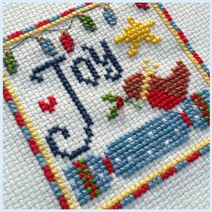 May include: Cross-stitch embroidery featuring the word "Joy" in blue thread, surrounded by a yellow, red, and blue border. A red and brown bird with a star above it is stitched in the bottom right corner.