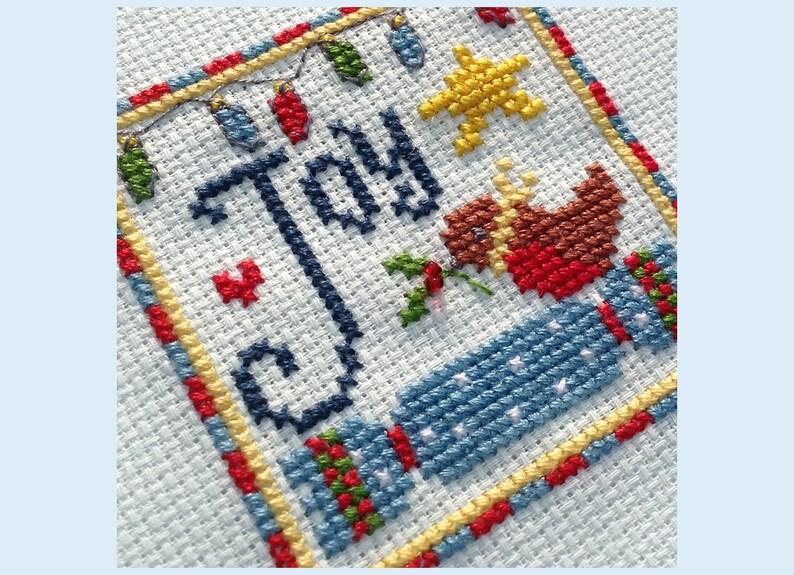 May include: Cross-stitch embroidery featuring the word "Joy" in blue thread, surrounded by a yellow, red, and blue border. A red and brown bird with a star above it is stitched in the bottom right corner.