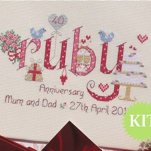 May include: Cross stitch kit featuring the word "ruby" in red with white polka dots. The letters are decorated with small embroidered images of hearts, birds, flowers, and a wedding cake. The text "Anniversary Mum and Dad 27th April 2014" is embroidered below the word "ruby". The kit is labelled "KIT" in a green circle.