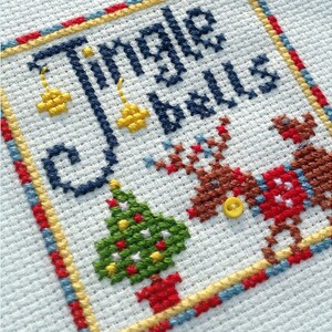 May include: Cross-stitch embroidery with the text "Jingle Bells" in blue thread. The design includes a green Christmas tree with red and yellow ornaments, two reindeer with red and brown accents, and yellow bells.