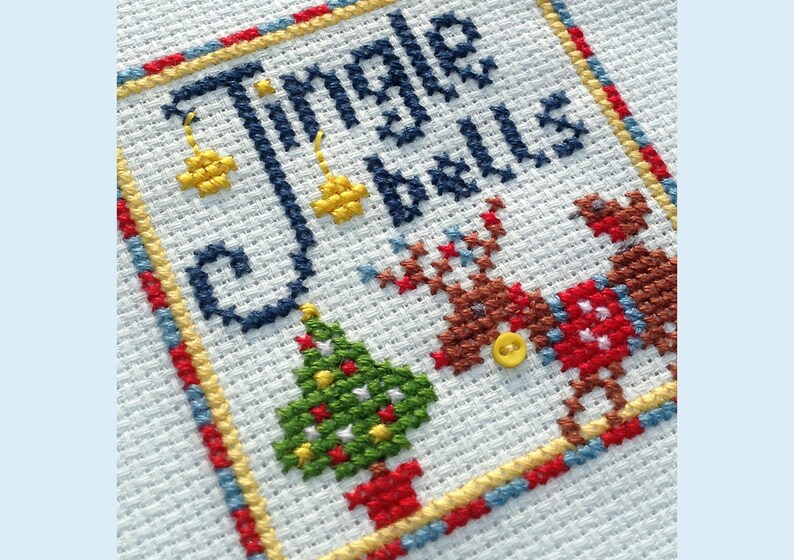 May include: Cross-stitch embroidery with the text "Jingle Bells" in blue thread. The design includes a green Christmas tree with red and yellow ornaments, two reindeer with red and brown accents, and yellow bells.