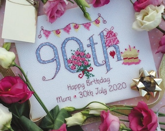 May include: A white cross-stitch embroidery with blue and pink lettering that reads "90th". The embroidery also includes a pink gift box, a bouquet of pink roses, and a pink and yellow birthday cake. The text below the number reads "Happy Birthday Mum - 30th July 2020".