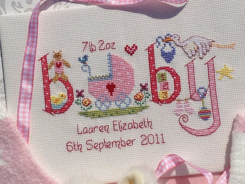 May include: Cross-stitch embroidery of the word "baby" in pink and red with a baby carriage, a duck, a teddy bear, a bird, a star, and a rattle. The text "7lb 2oz Lauren Elizabeth 6th September 2011" is below the word "baby".