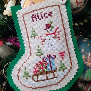May include: A green felt stocking with a white embroidered design of a snowman with a red hat and scarf, a sled with presents, and trees. The stocking has the name "Alice" embroidered on it.