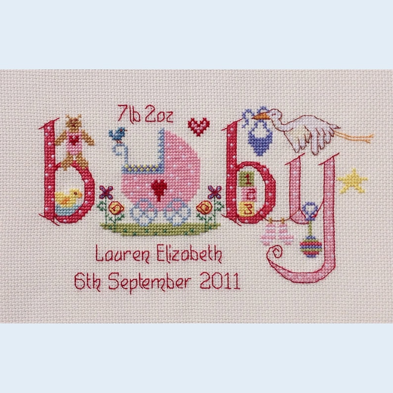 May include: A pink and white cross-stitch embroidery with the word "baby" spelled out in large letters. The letters are decorated with various baby-themed images, including a baby carriage, a teddy bear, a bird, a stork, and a rattle. The text "7lb 2oz" is above the word "baby" and the text "Lauren Elizabeth 6th September 2011" is below the word "baby".