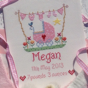 May include: A pink and white cross-stitch embroidery with a baby carriage, flowers, and a star. The text reads "Megan, 11th May 2013, 7 pounds 3 ounces".