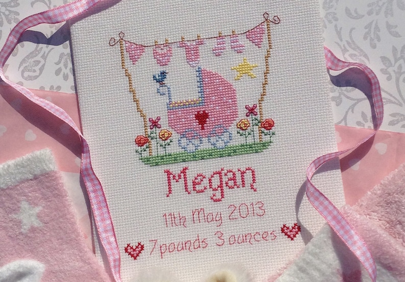 May include: A pink and white cross-stitch embroidery with a baby carriage, flowers, and a star. The text reads "Megan, 11th May 2013, 7 pounds 3 ounces".