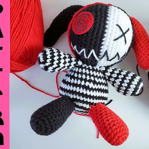 May include: A black and white crocheted amigurumi toy with red accents. The toy has a stitched-on face with a red swirl and an X for eyes. The toy is sitting on a white surface with red and black yarn balls.