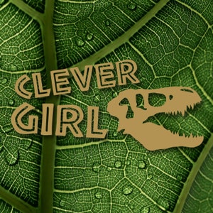 May include: A green leaf with a brown dinosaur skull and the text "CLEVER GIRL" in brown outlined letters.