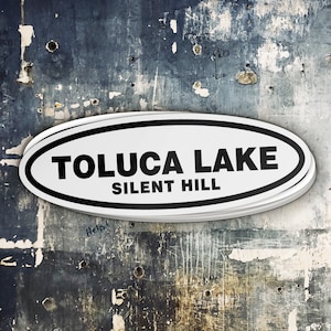 Puede incluir: Una pegatina ovalada blanca con texto negro que dice "TOLUCA LAKE SILENT HILL" sobre un fondo azul y gris texturizado.