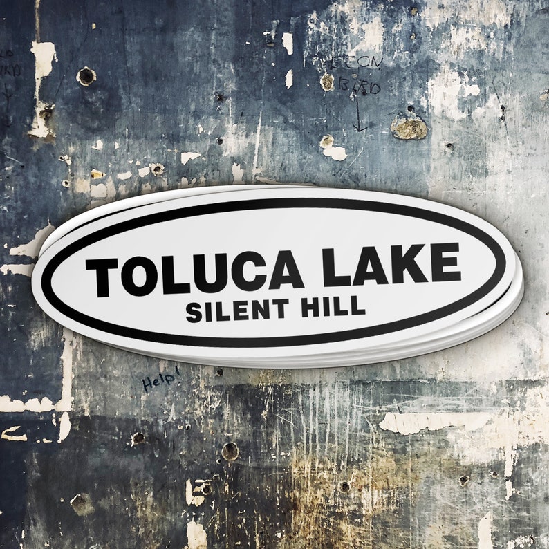 Puede incluir: Una pegatina ovalada blanca con texto negro que dice "TOLUCA LAKE SILENT HILL" sobre un fondo azul y gris texturizado.