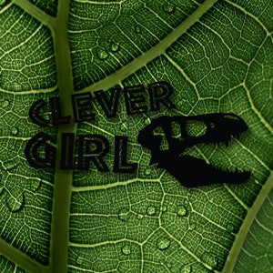 May include: A green leaf with water droplets on it. The words "CLEVER GIRL" are printed in black on the leaf, along with a silhouette of a dinosaur skull.