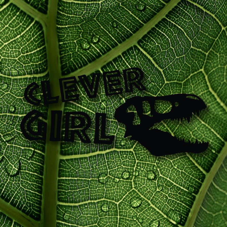 May include: A green leaf with water droplets on it. The words "CLEVER GIRL" are printed in black on the leaf, along with a silhouette of a dinosaur skull.