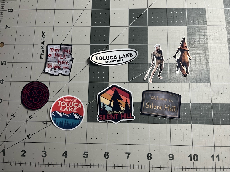 Puede incluir: Una colecci&oacute;n de nueve pegatinas con varios dise&ntilde;os de la serie de videojuegos Silent Hill. Las pegatinas incluyen un s&iacute;mbolo rojo y negro, una escena de monta&ntilde;a de estilo retro con una silueta de monstruo, un cartel que dice "Bienvenido a Silent Hill" y una pegatina con el texto "Silent Hill Toluca Lake".