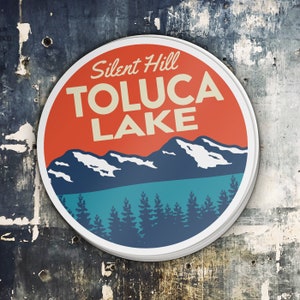 Puede incluir: Una pegatina redonda con un dise&ntilde;o rojo, blanco y azul. La pegatina presenta una cordillera con picos cubiertos de nieve, un lago azul y un bosque de &aacute;rboles de hoja perenne. El texto "Silent Hill Toluca Lake" est&aacute; impreso en una fuente de estilo vintage.