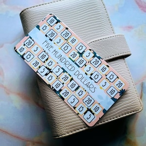 May include: A beige textured leather-like personal planner with a strap closure, featuring a rectangular savings tracker card on top. The card has a floral background and displays the text FIVE HUNDRED DOLLARS with various numerical values.