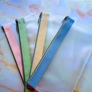 May include: A collection of clear, zippered pouches with colorful accents. Each pouch has a different colored zipper pull: pink, green, yellow, and blue. The pouches are arranged in a fan shape, showcasing their contents.