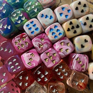 May include: A collection of square dice featuring marbled, translucent, and solid finishes in various colors. Each die displays a unique arrangement of heart-shaped pips in metallic gold, silver, or black, replacing traditional numbered dots.