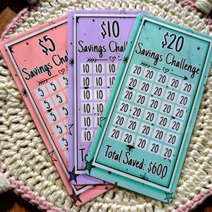 Peut inclure: Trois cartes color&eacute;es sur le th&egrave;me du d&eacute;fi d'&eacute;pargne. Les cartes sont rose, violette et turquoise. Chaque carte a un montant diff&eacute;rent&nbsp;: 5&nbsp;$, 10&nbsp;$ et 20&nbsp;$. Les cartes comportent une grille de nombres pour suivre les &eacute;conomies. La carte turquoise affiche le texte&nbsp;&laquo;&nbsp;Total &eacute;conomis&eacute;&nbsp;: 600&nbsp;$&nbsp;&raquo;.