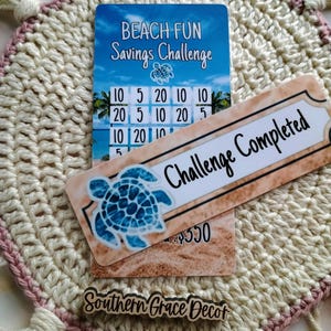 May include: A beach-themed savings challenge card with a grid of numbers and a sea turtle design. The card has a bookmark attached that says "Challenge Completed".