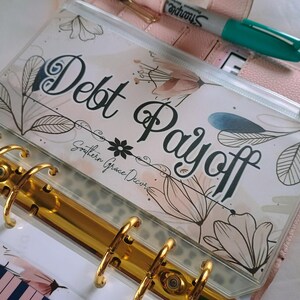 Op de afbeelding: Een doorzichtige plastic ritszak met een bloemenontwerp en de tekst "Debt Payoff" en "Southern Grace Decor". De zak zit in een gouden map.
