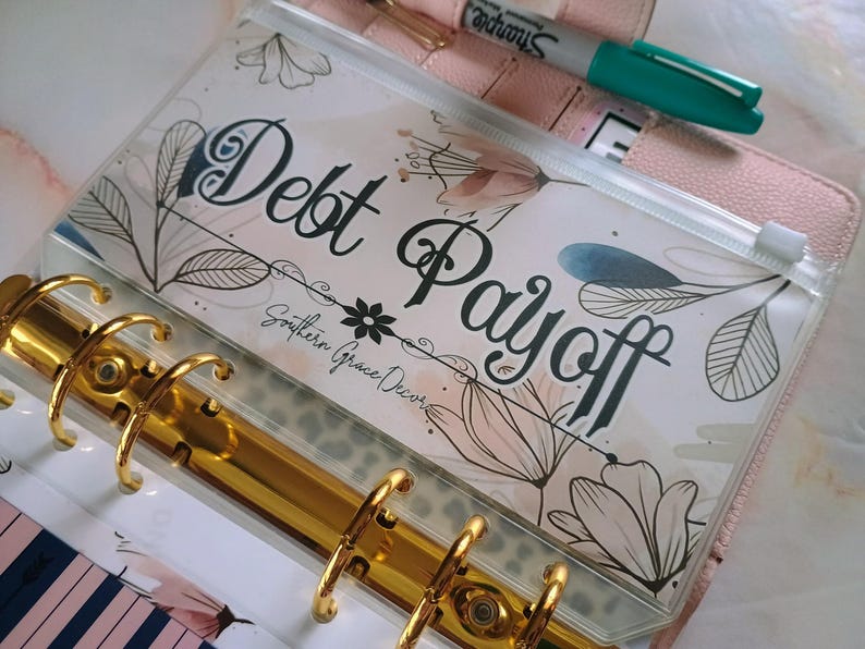 Op de afbeelding: Een doorzichtige plastic ritszak met een bloemenontwerp en de tekst "Debt Payoff" en "Southern Grace Decor". De zak zit in een gouden map.