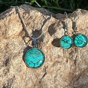 May include: A jewelry set featuring a pendant necklace and matching earrings. The pendant and earrings have a round, silver-toned frame with a teal and black marbled design. The necklace has a silver chain.