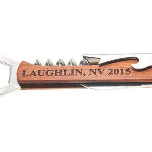 May include: A wooden wine opener with a silver bottle opener, corkscrew, and foil cutter. The handle is engraved with the text "Laughlin, NV 2015".