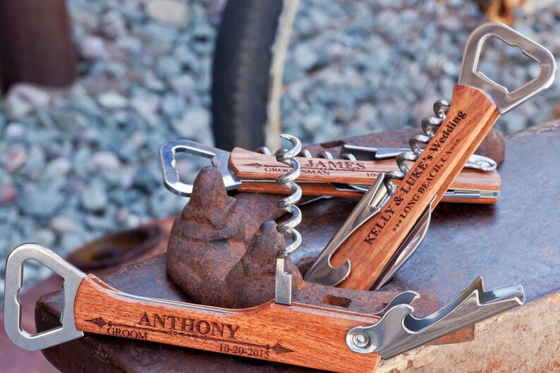 May include: Three wooden bottle openers with engraved text. The top opener says "Kelly & Luke's Wedding...Long Beach, CA." The middle opener says "James, Groomsman." The bottom opener says "Anthony, Groom 10-20-2015."