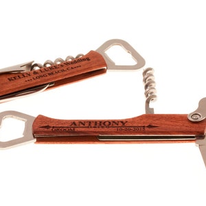 May include: Two wooden multi-tools with engraved text. The top tool has a bottle opener, corkscrew, and a small knife. The bottom tool has a bottle opener, a corkscrew, and two small knives. The top tool is engraved with "KELLY & LUKE'S Wedding *** LONG BEACH, CA ***". The bottom tool is engraved with "ANTHONY GROOM 10-20-2015".