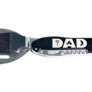 May include: Black multi-tool with a spatula, brush, and bottle opener. The tool has the word "DAD" engraved on it with a flame and spatula icon.