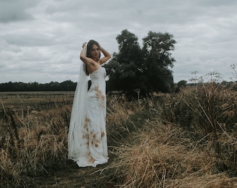 May include: A woman in a white wedding dress with floral embroidery stands in a field of tall grass. She is wearing a veil and has her hair pulled back. The sky is overcast and there are trees in the background.