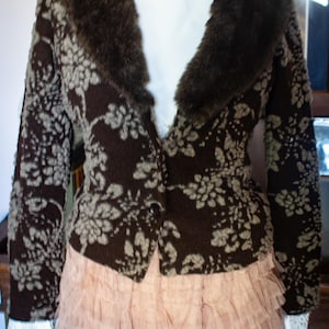 Könnte beinhalten: Eine dunkelbraune Jacke mit hellgrauem Blumenmuster, einem dunkelbraunen Kunstfellkragen und einem Knopfverschluss. Die Jacke wird über einem weißen Oberteil mit Spitzenbündchen und einem mehrlagigen, hellrosa Tüllrock getragen.