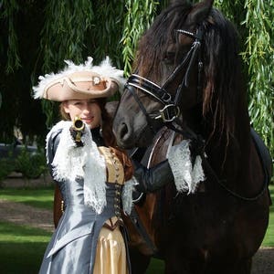 May include: A woman in a gray and gold dress and a feathered hat holds a pistol and stands next to a black horse wearing a bridle.