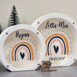 May include: Two white wooden piggy banks with rainbow designs and personalized names. The larger bank says "Lotta-Mia" and has the date "06.01.2017". The smaller bank says "Brynn".
