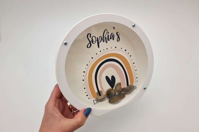 May include: White wooden piggy bank with a clear plastic window. The window shows a rainbow design with a heart and the text "Sophia's Dreamcatcher Wishes".