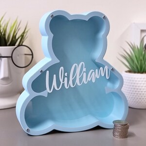 May include: A light blue wooden money box shaped like a bear with the name "William" cut out in white on the front. The money box is sitting on a grey surface with a stack of coins in front of it.