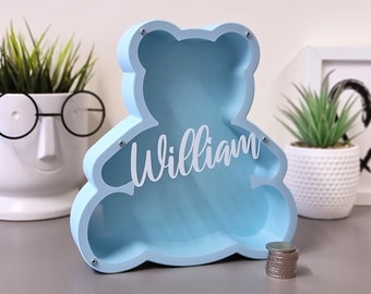 May include: A light blue wooden piggy bank shaped like a bear with the name "William" cut out in white on the front. The piggy bank is sitting on a gray surface with a stack of coins in front of it.