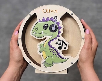 May include: A wooden coin bank shaped like a circle with a dinosaur wearing headphones and musical notes. The bank is personalized with the name "Oliver".