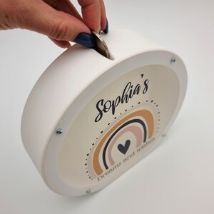 May include: White ceramic piggy bank with a slot on top. The front features a rainbow design with a heart in the center and the text "Sophia's Dreams and Wishes".