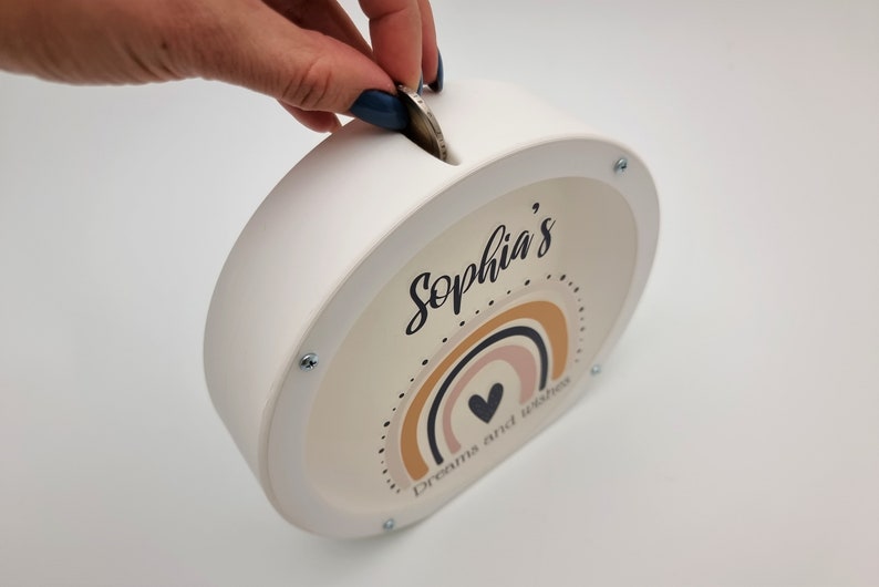 May include: White ceramic piggy bank with a slot on top. The front features a rainbow design with a heart in the center and the text "Sophia's Dreams and Wishes".