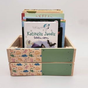 May include: A wooden crate painted green and decorated with a dinosaur pattern, filled with a stack of children's books. The top book is titled "Katinelis Juodis Saldziu sapnu" in Lithuanian.