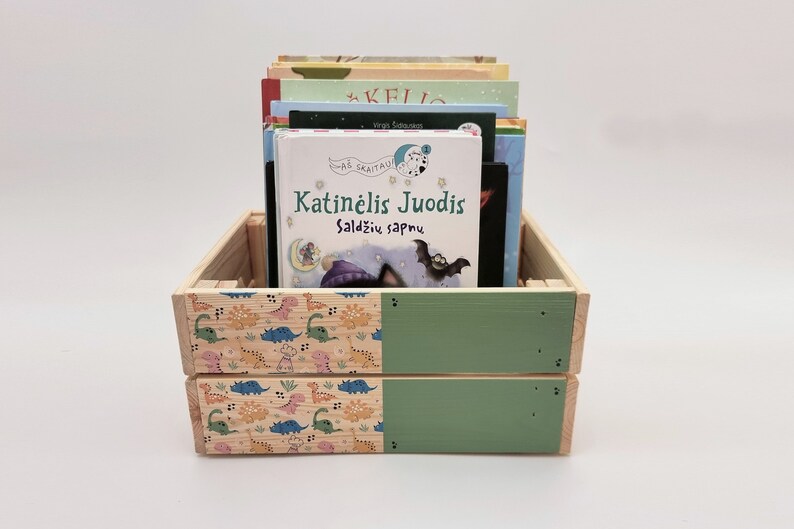 May include: A wooden crate painted green and decorated with a dinosaur pattern, filled with a stack of children's books. The top book is titled "Katinelis Juodis Saldziu sapnu" in Lithuanian.