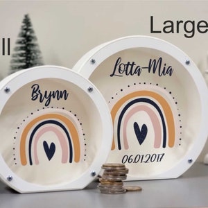 May include: Two white wooden piggy banks with rainbow designs and a heart. The larger bank says "Lotta-Mia 06.01.2017" and the smaller bank says "Brynn".