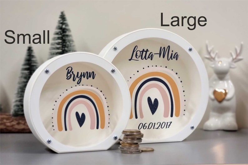 May include: Two white wooden piggy banks with rainbow designs and a heart. The larger bank says "Lotta-Mia 06.01.2017" and the smaller bank says "Brynn".