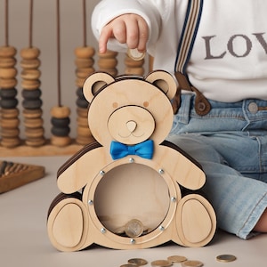 May include: A wooden teddy bear money box with a blue bow tie and a clear plastic coin slot. A child is putting a coin into the money box.