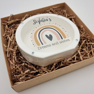 May include: A white ceramic coin bank with a rainbow design and the text "Sophia's Dreams and Wishes". The coin bank is in a brown cardboard box with brown shredded paper.