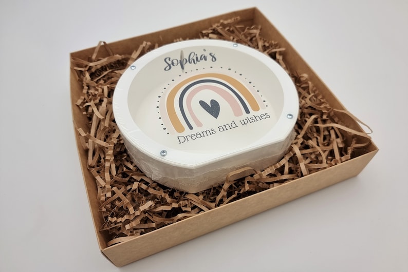 May include: A white ceramic coin bank with a rainbow design and the text "Sophia's Dreams and Wishes". The coin bank is in a brown cardboard box with brown shredded paper.