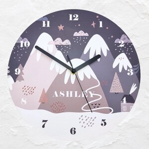 May include: A round, purple and pink wall clock with a snowy mountain scene and the name "ASHLEY" printed on the face. The clock has black hands and white numbers.